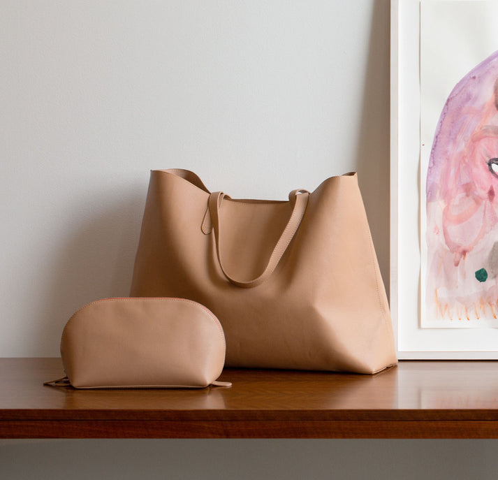 Beige tote bag and pouch on a wooden table beside a framed abstract painting.